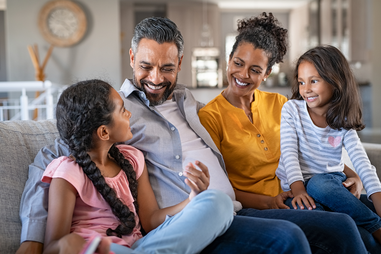 Cheerful multiethnic family at home sitting on sofa listening to daughter. Happy couple with two girls relaxing at home together. Smiling indian parents talking to their daughter in the living room.